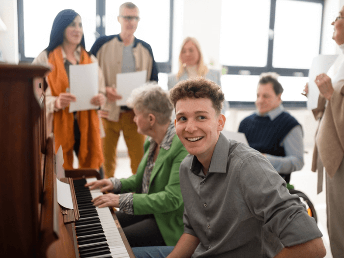 A group of diverse people of differing ages surrounding a piano singing while two play. Representing community music groups.