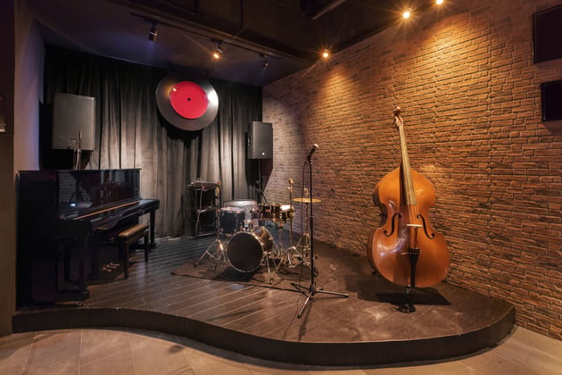 Photograph of an empty stage in a jazz bar. Unattended instruments are set up, a double bass, a piano and a drum kit.