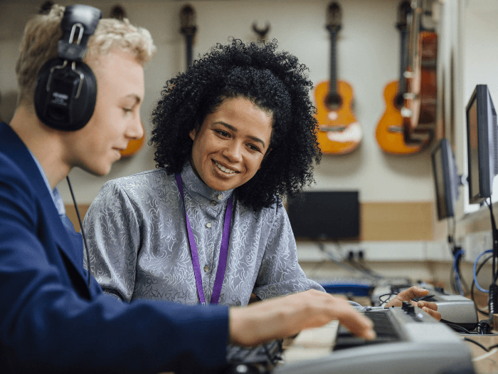 Female teacher is sitting with one of her students in a music lesson at school. He is learning to play the keyboard and is wearing headphones.