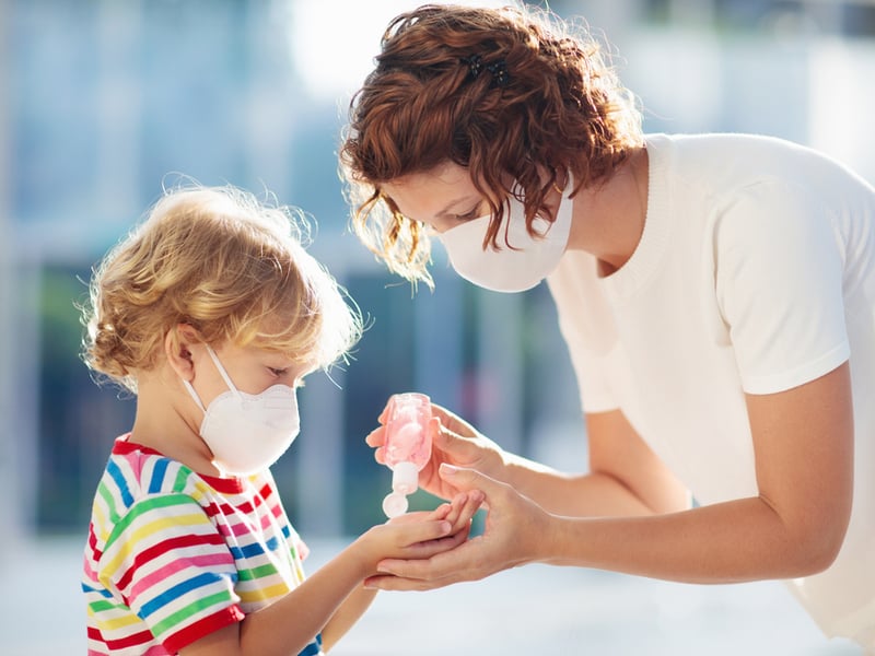 Woman applies hand sanitizer to a child wearing a facemask and a rainbow coloured t-shirt.