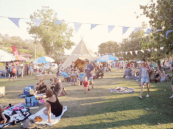 Blurred, defocused background of people attending a fair/summer festival, with tents, lights. A family on a picnic blanket is in the foreground.
