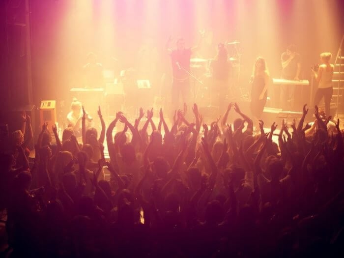 Crowd with hands raised in a small venue watching a band on stage, the image is cast in a pale yellow glow from the stage lights.