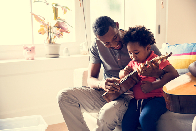 Photograph of a man sat with a child, playing ukulele in what appears to be a home setting with white walls and a house plant.