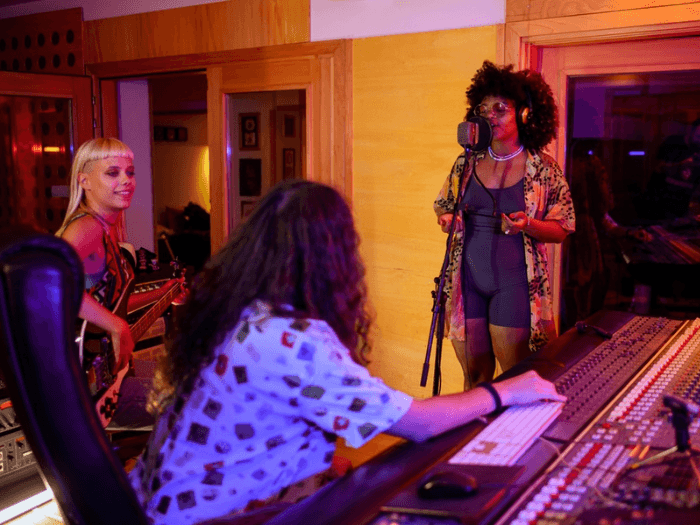Two female session musicians in the studio, one playing bass and the other singing into a microphone. The sound technician is sitting at the mixing desk, turned towards them.