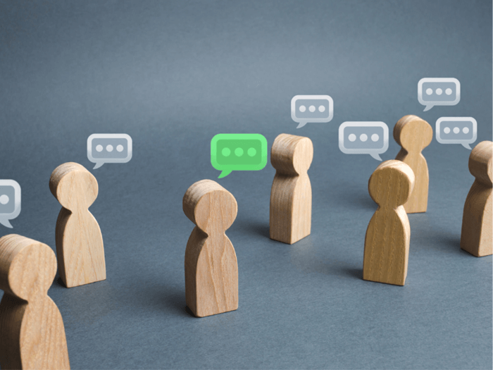 7 wooden shaped non gender figures on grey surface, with 3 dots in a speech bubble in white, one of the figures has a speech bubble in green showing they are talking.