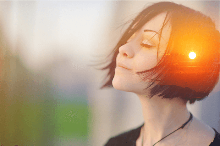 Women with short dark hair, her eyes are closed and she is breathing deeply, an orange sunrise is subtly imposed over the head to symbolise mindfulness and new beginnings.