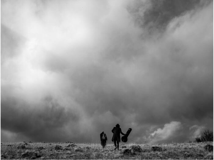 Black and grey image of two people, one holding a guitar case, walking up a hill together with a stormy sky above.