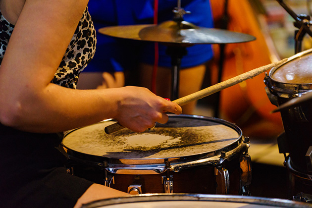 A person is playing on a drum kit, the photograph just shows us a close up of the kit, one well used stick, and the arm and shoulder of the person who is wearing a leopard print vest.