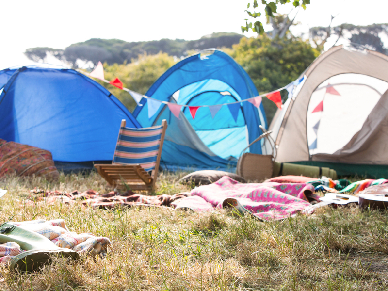 Photograph of a sunny festival camping scene, blankers, chairs and tents are strewn around.