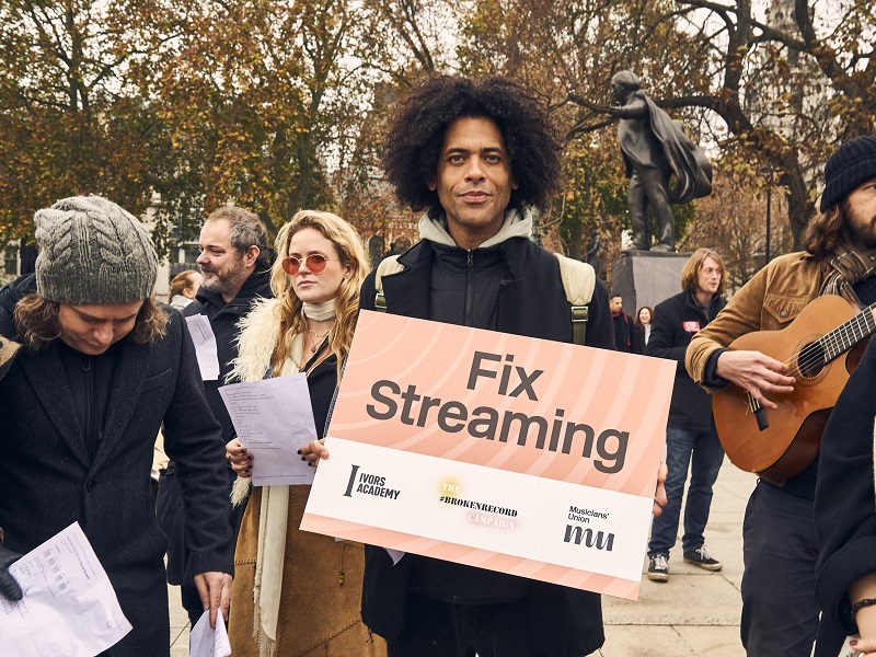 People at a protest with Autumn trees in the background, man in foreground is holding a fix streaming campaign board