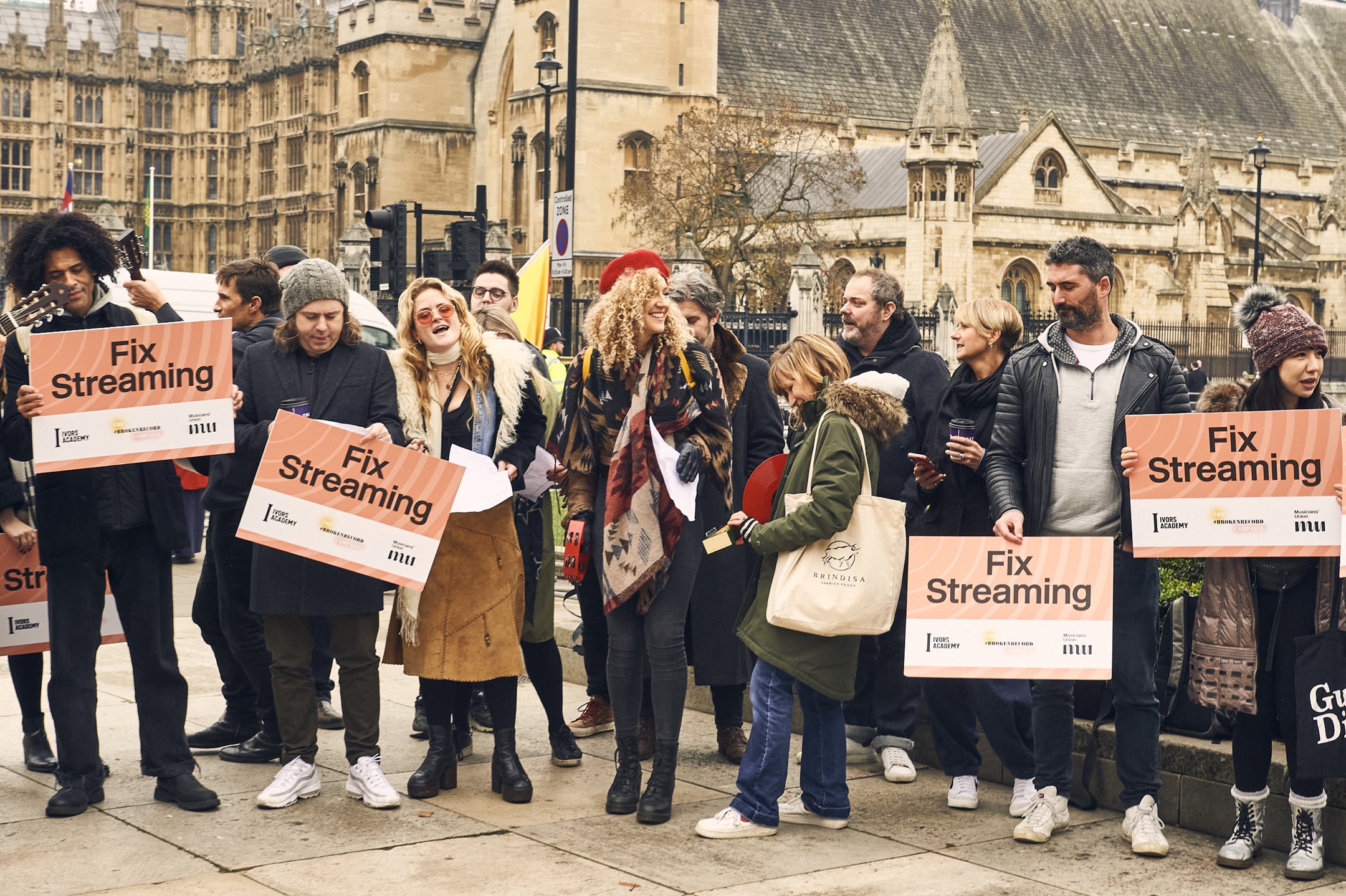 Photograph of a large group of people standing outside the houses of parliament, some appear to be singing and some are holding placards reading 