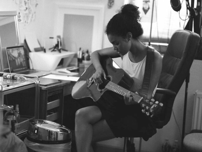 Black and white image of young woman sat in front of a laptop playing a guitar resting on her knee.