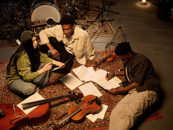Three young musicians, collaborating on music composition, sitting on a carpet with string instruments and sheet music discussing ideas.