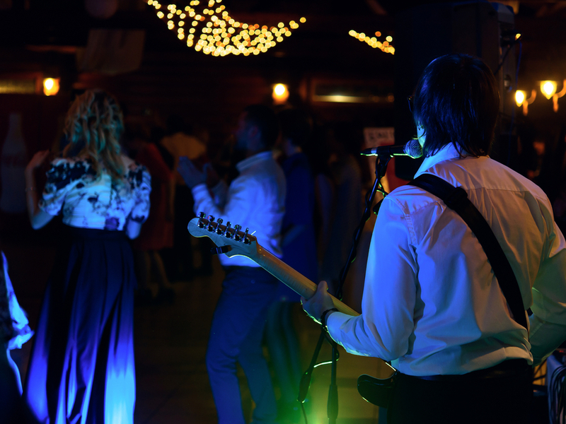 Photograph of a function band performing at a wedding, as seen from behind. We see the back of a guitarist in blue lighting.