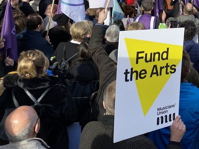 A fund the arts sign from the Musicians' Union is held aloft in a crowd