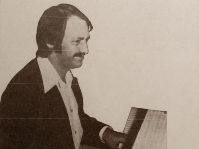 Black and white newspaper clipping image of Gerry smiling sat a piano.