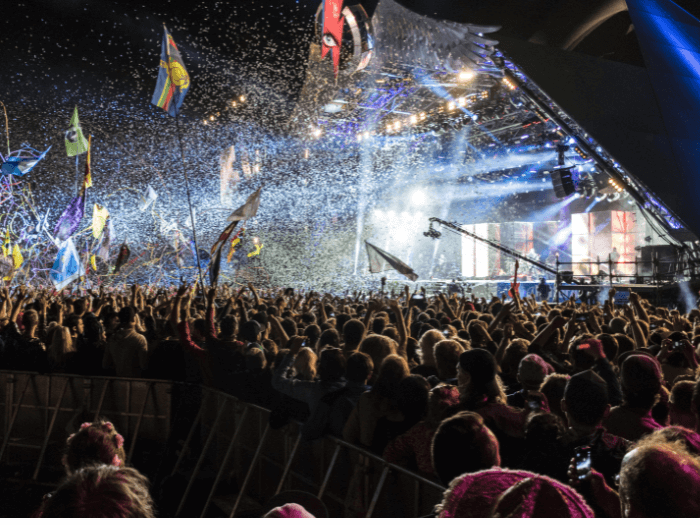 A crowd of people at night watching the Pyramid stage at Glastonbury festival, streamers, confetti and flags are in the air.