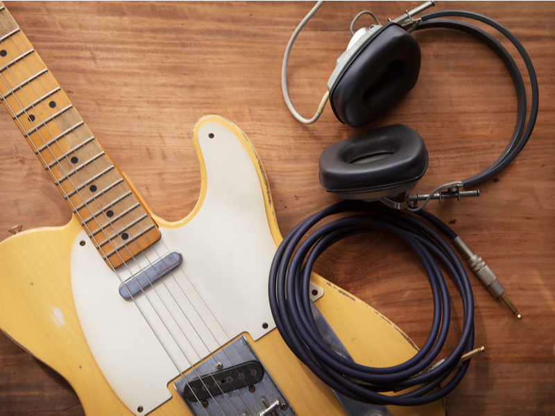 Photograph of a sandy coloured electric guitar laid out on a wood table with a loop of amplifier cable and earphones also displayed.