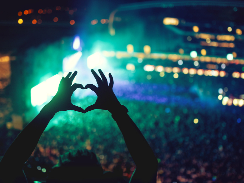 Silhouette of a person holding up their hands in a heart shape, against a stadium audience at a live music event.