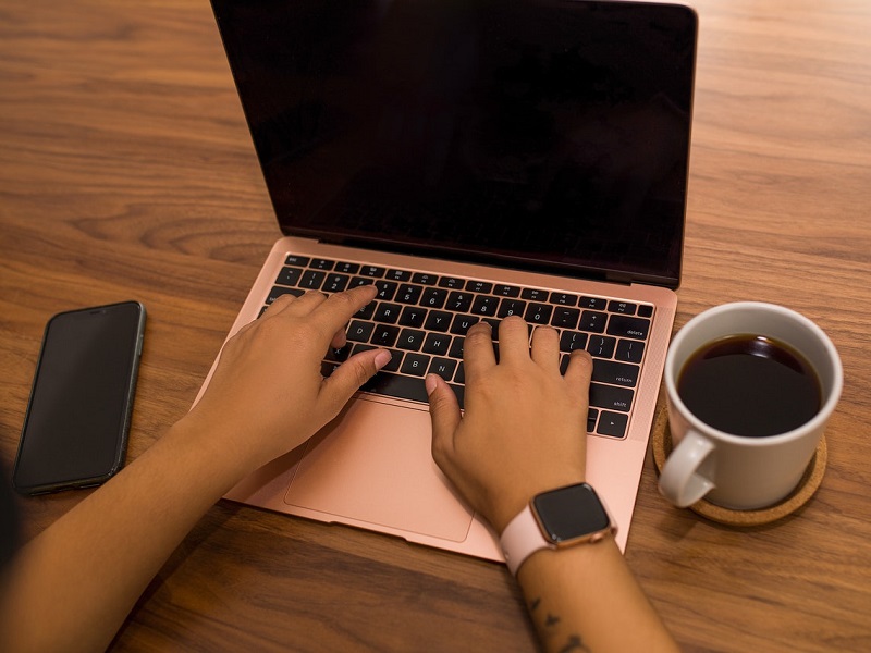An adults two hands and arms are typing on a rose gold laptop, the screen content is not visible. They also have a cup of coffee and a phone on the table.