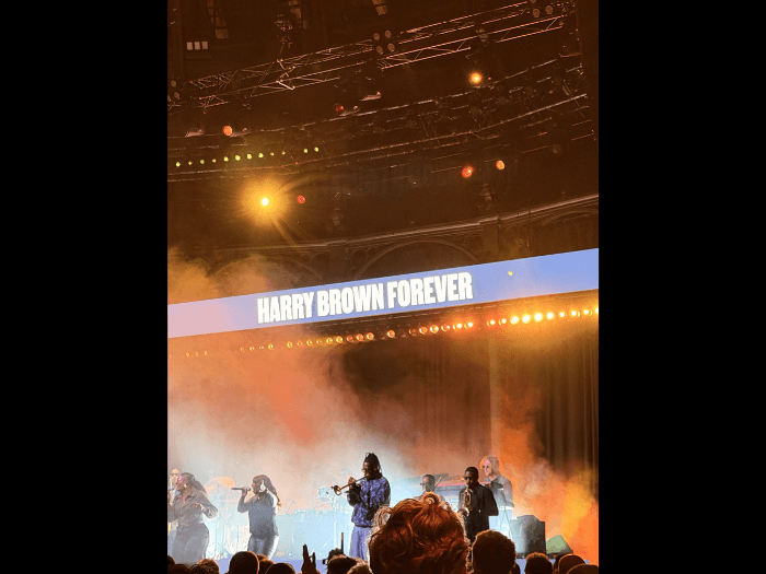 Ezra Collective's tribute at a recent gig, they are performing on stage with a large blue sign that says “Harry Brown Forever”.