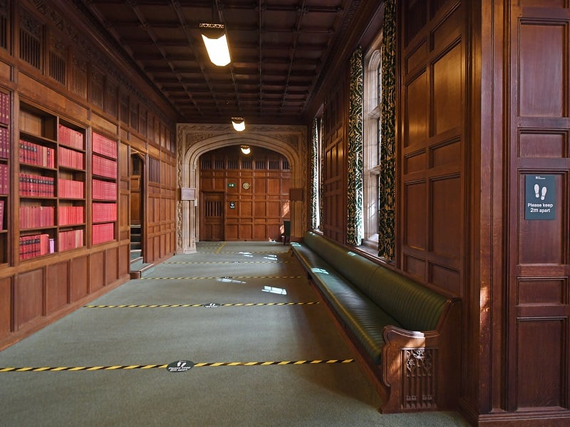 Photograph of a corridor inside the Houses of Parliament, striped tape on the floor indicates how people must stand 2 metres appart to socially distance.