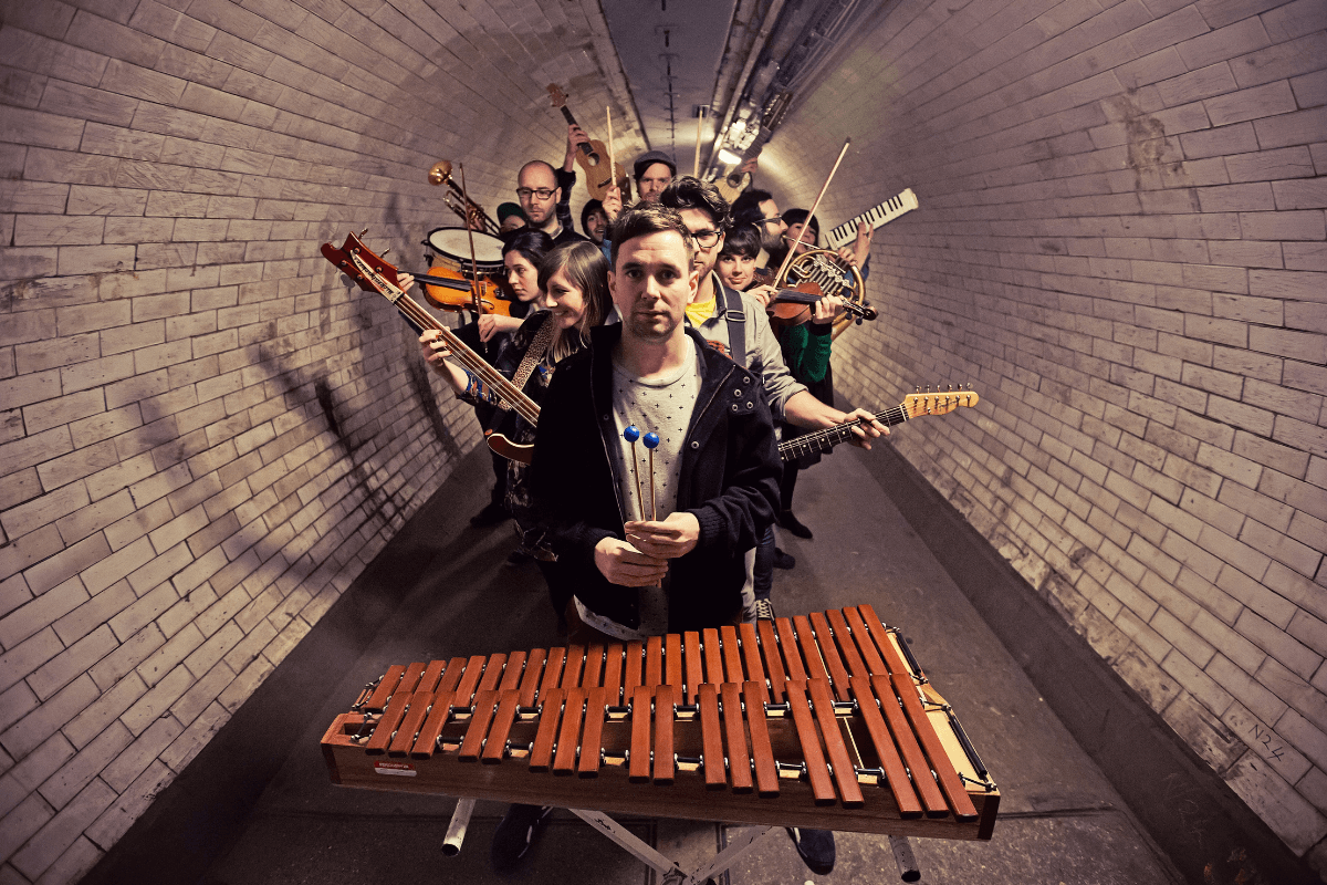 Press shot of Paul Russell and his orchestral ensemble Human Pyramids, holding their instruments in a tunnel.