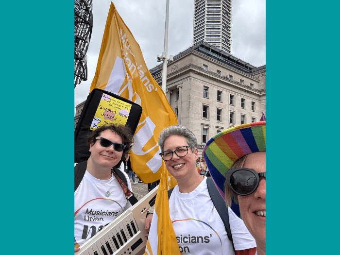 Jenny is wearing a rainbow hat and two MU members are wearing Union branded t-shirts, carrying flags and a keyboard.