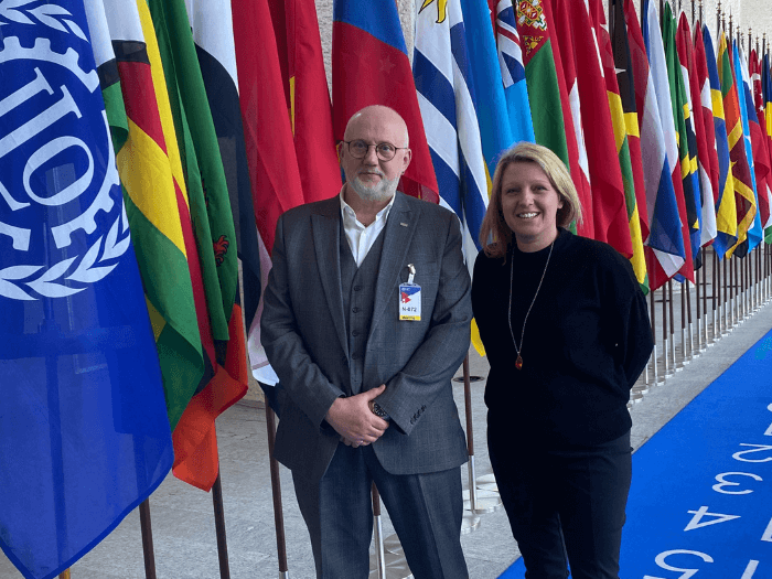 Naomi Pohl and Dave Webster stand together in front of row of flags of member states attending the ILO.