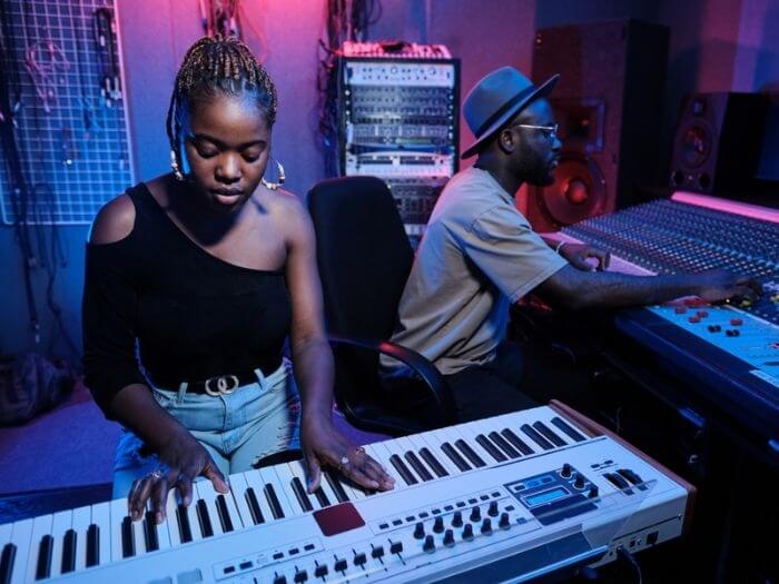 Two Black musicians in the recording studio using the mixing desk and playing keyboard.