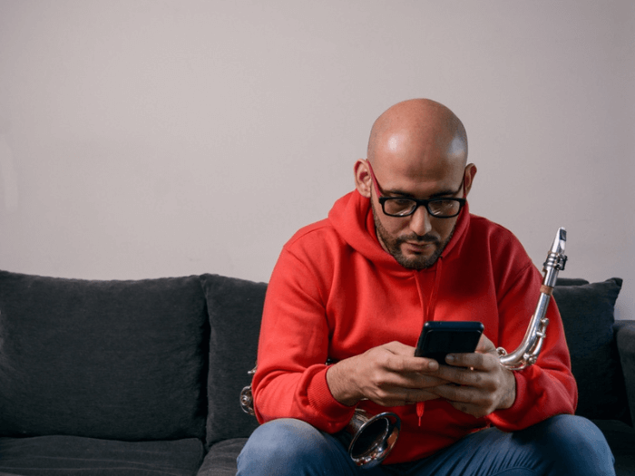 Middle-aged man sitting on a sofa with a saxophone on his legs, browsing on his phone.