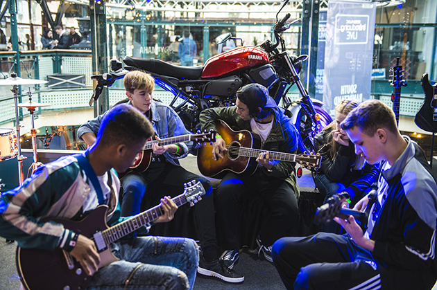 Photograph of five young people sat in a circle playing guitars.