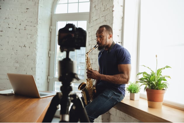 Photograph of a musician playing saxophone at home, playing into a webcam that is set up to record.