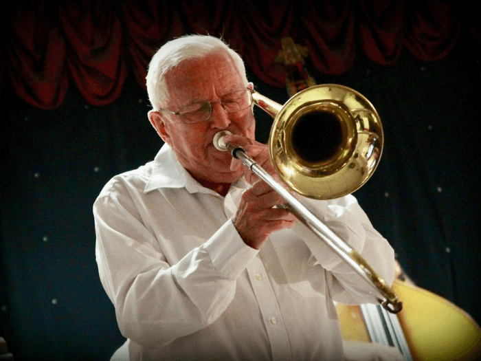 Jack onstage as an older man, wearing a white shirt and playing trombone.