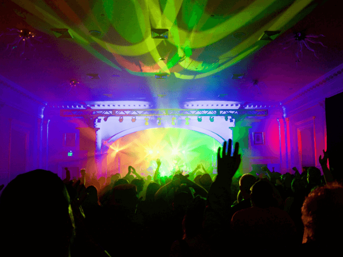 Inside a small dark venue with vibrant rainbow lights, the crowd and band on stage are in silhouette.
