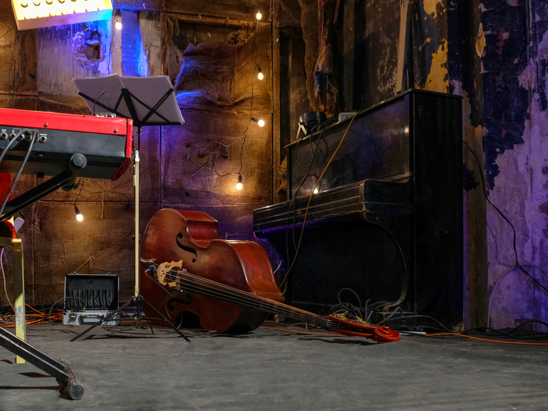 Photograph of a number of instruments left backstage, a well aged piano, an upright bass lying on its side, a red synth on a stand and a music stand, as well as fairy lights and wires and visible.