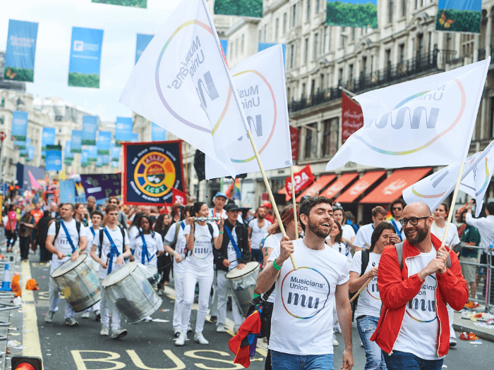 John Shortell wearing a Musicians' Union t-shirt with rainbow logo, holding a flag, during a Pride march in 2023. He is joined by colleagues and other members who are performing as they walk.
