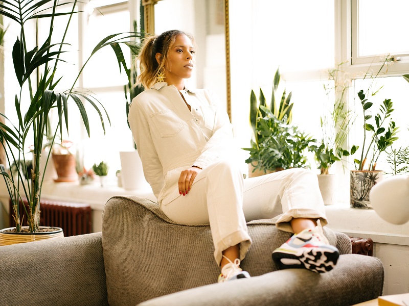 A photograph of Kelli-Leigh, she's sitting on a sofa in a bright, plant filled room, looking into the space behind the camera thoughtfully.
