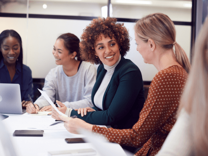 Group of diverse women sat talking around a table, making notes with paper and laptops, smiling as they network and share ideas.