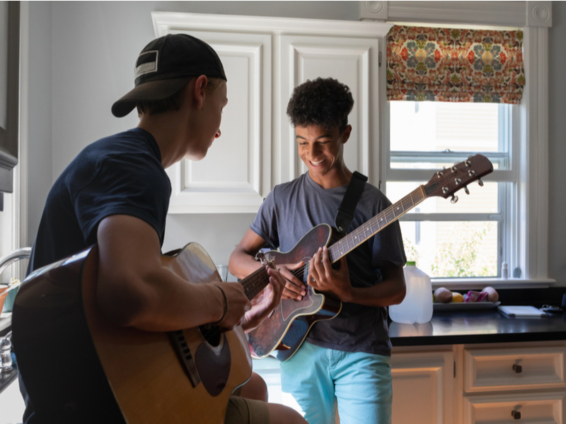 Two young adults playing their guitars in the kicthen, both are holding acoustic guitars as if they are preparing to play them.
