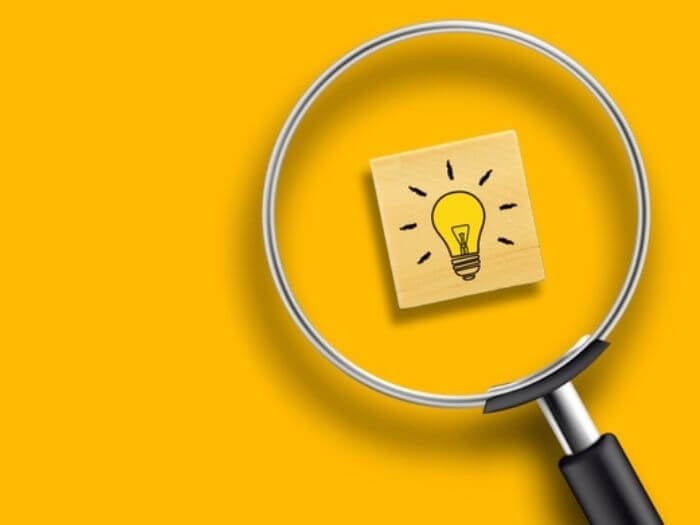 Magnifying glass over a light bulb icon on a wooden block, with yellow background.