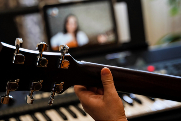 Photograph of the back of a guitar neck, with a hand in a playing position. In the background there appears to be a teacher on an i-pad giving an online music lesson.