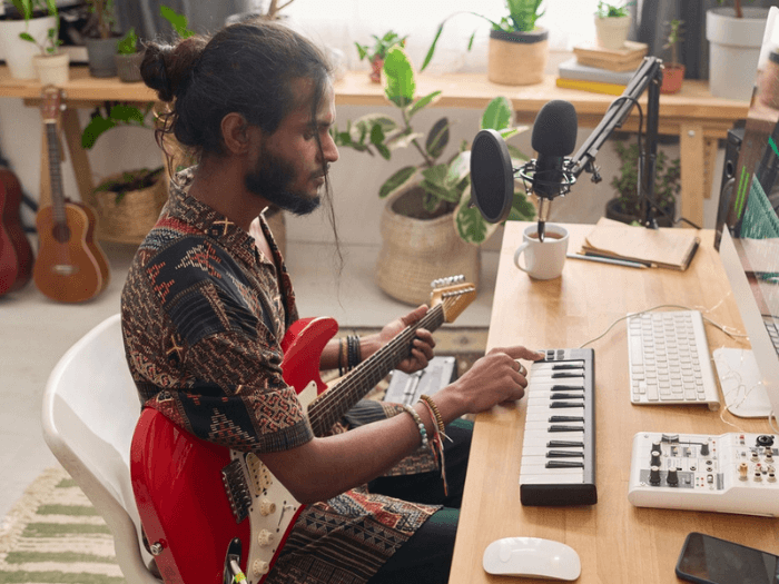A South Asian musician holding an electric guitar and playing a midi keyboard while sat at a desk.