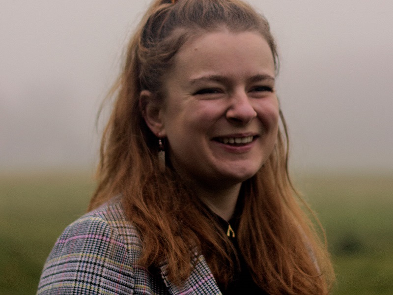 Photograph of musician Lizzy Hardingham, she is laughing and looking past the camera in a brightly coloured jacket,