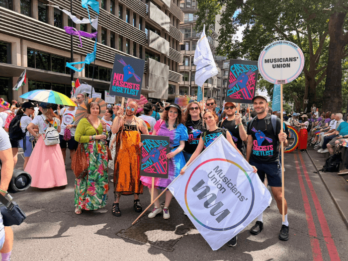 The MU delegation en route, holding flags with the union logo in a rainbow circle on a white background, and pink, teal, blue and black placards that say 
