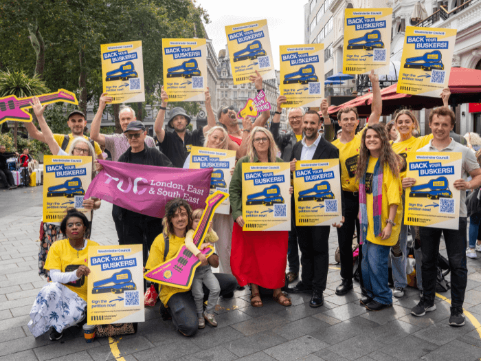 Group of buskers, supporters, and MU staff outside at the site of the Leicester Square busking pitch. Some are wearing t-shirts, or holding posters and props, that say ‘back your buskers’.