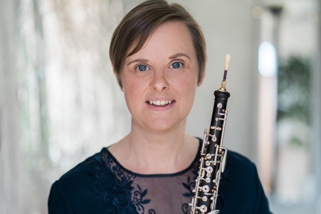 Photograph of Louise, wearing a dark, professional looking shirt and smiling whilst holding a wind instrument. The background is white, bright and sunny.