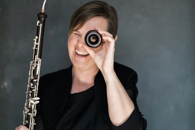 Photograph of a smiling woman, holding her reed instrument and looking through a detached part of it with one eye.