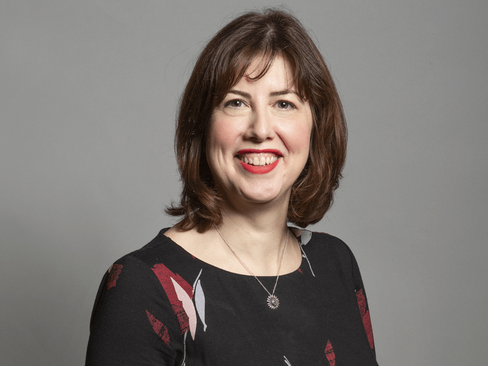 Official Gov portrait of Lucy Powell MP smiling. Cropped headshot to shoulders with grey background.