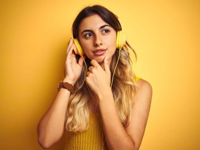 Young woman deep in thought wearing yellow headphones against a warm yellow background.